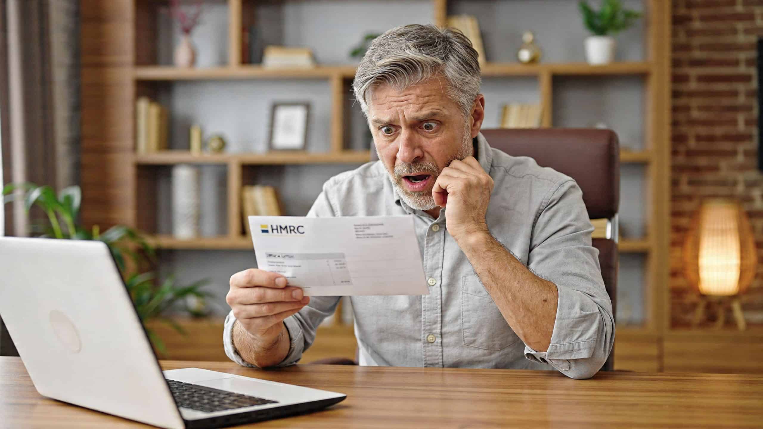 A middle-aged man looking shocked and concerned while holding an HMRC tax letter at his desk.