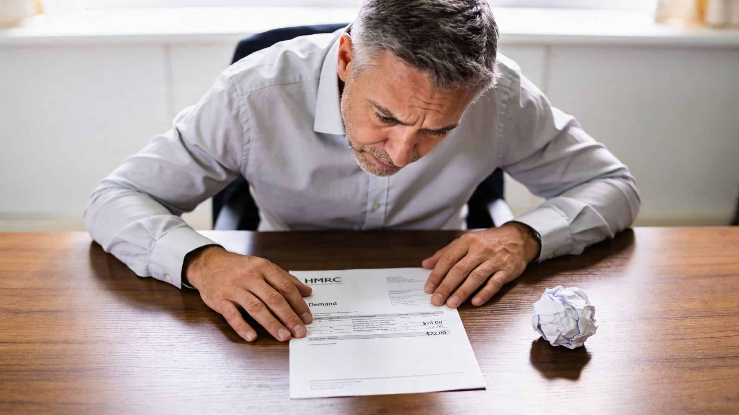 A stressed man sitting at a wooden desk reading a tax demand letter from HMRC.